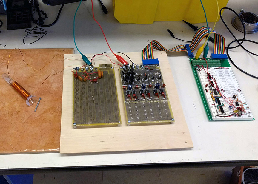 The induction heater setup on a lab bench. A wire coil is wrapped around a glass graduated cylinder which is connected to two circuit boards and a breadboard.