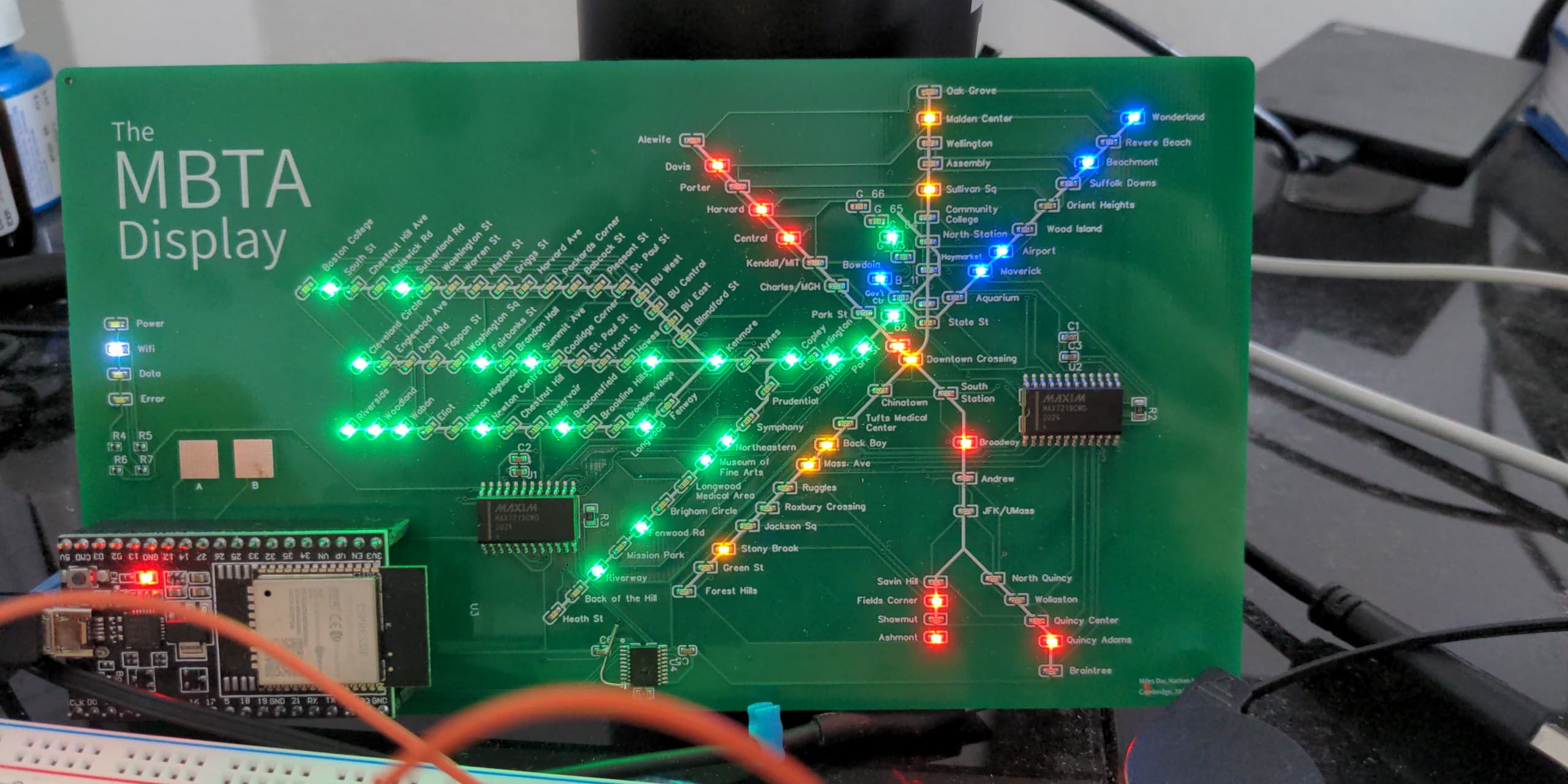 A green PCB with the text "The MBTA Display" in the upper left. The PCB has a map of the Boston MBTA with LEDs at each stop. Some LEDs are lit.